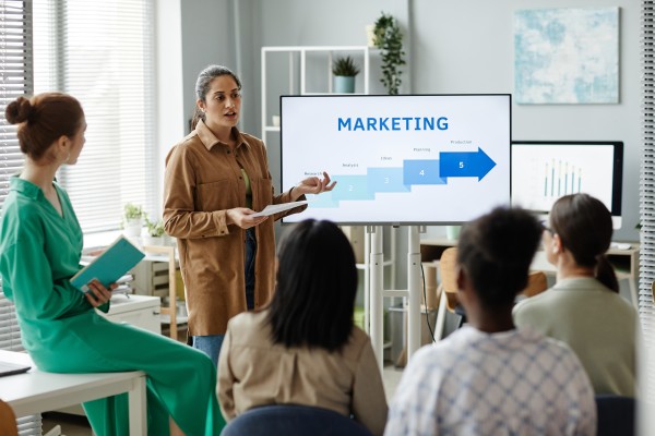 Woman presenting a five-step MARKETING arrow on a screen to seated colleagues in an office meeting.