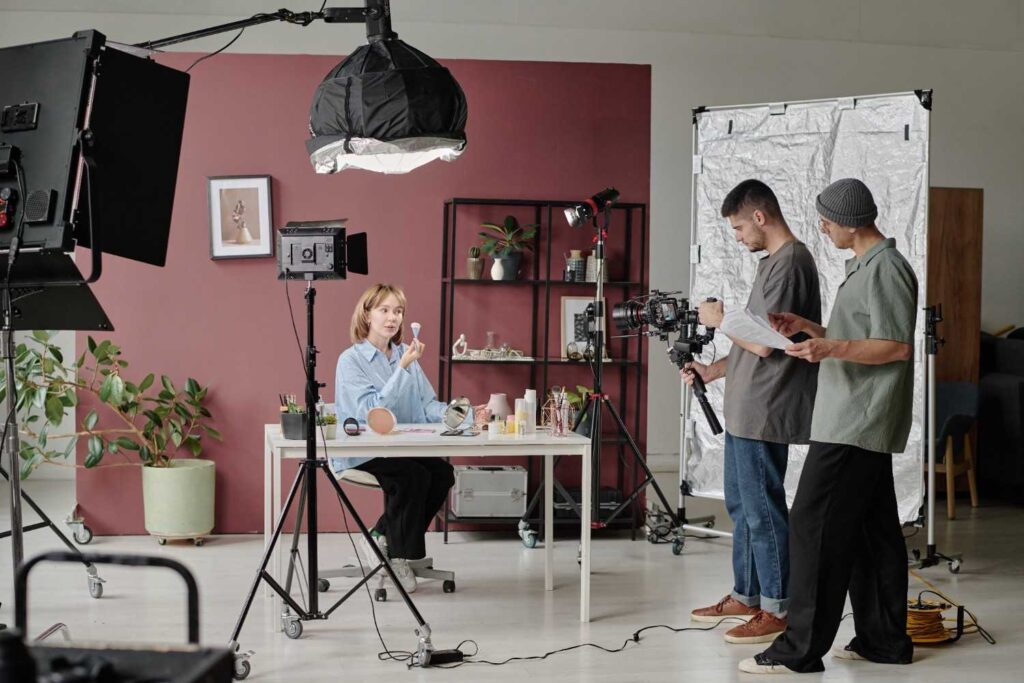 Woman at desk demonstrating makeup on set with lights, camera and two crew members consulting a script.