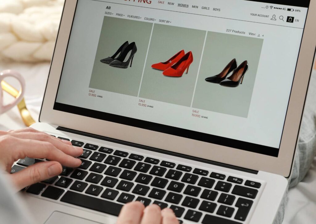 Hands on a laptop showing an online shop with three women's high heels: studded black, red and black patent.