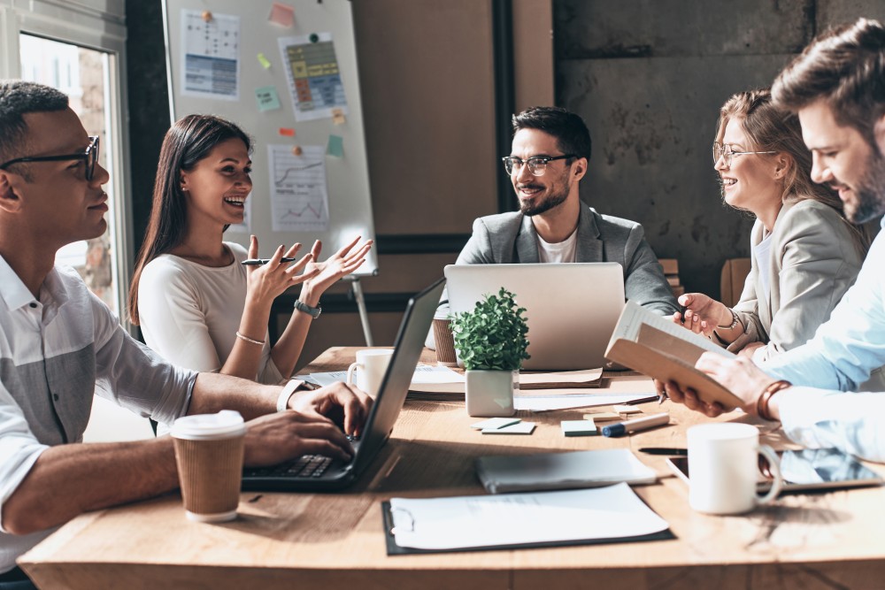 Five colleagues at a meeting table with laptops and coffee as a woman gestures while the team reviews flipchart notes.