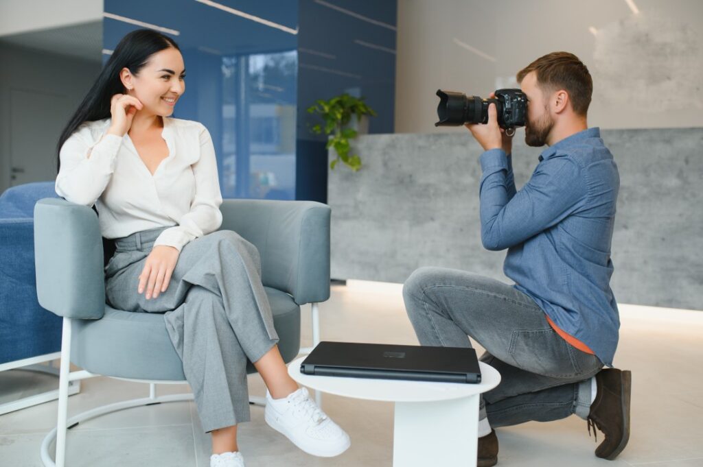 Smiling woman in white blouse and grey trousers sits in an armchair as a kneeling photographer takes her portrait with a DSLR.