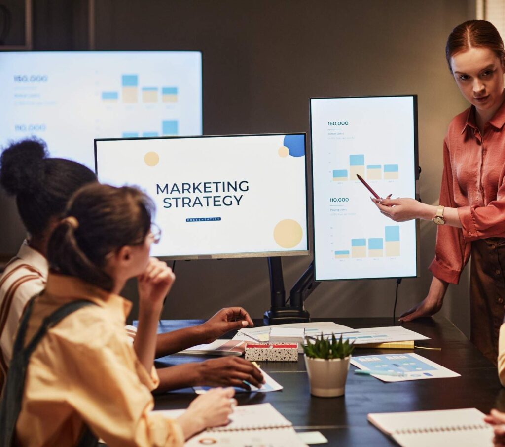 Team meeting in a conference room with a presenter pointing at a vertical screen of bar charts beside a monitor titled Marketing Strategy