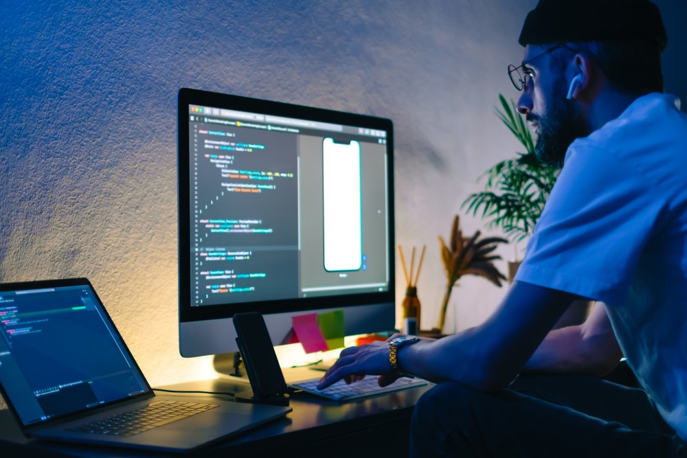 Developer in beanie and glasses coding at a desk, desktop shows a phone app mockup while laptop displays code.