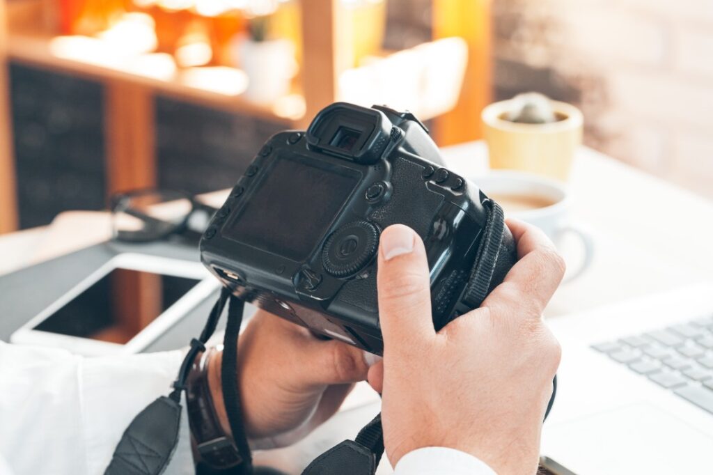 Person's hands holding a DSLR camera, checking its rear LCD on a desk with coffee, tablet and laptop.