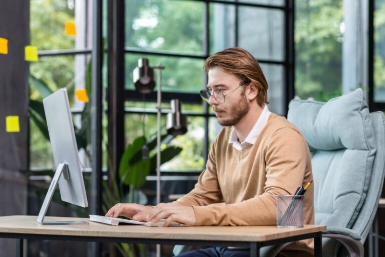 Young man with glasses and beard typing at a desktop computer in a modern office with large windows and sticky notes.